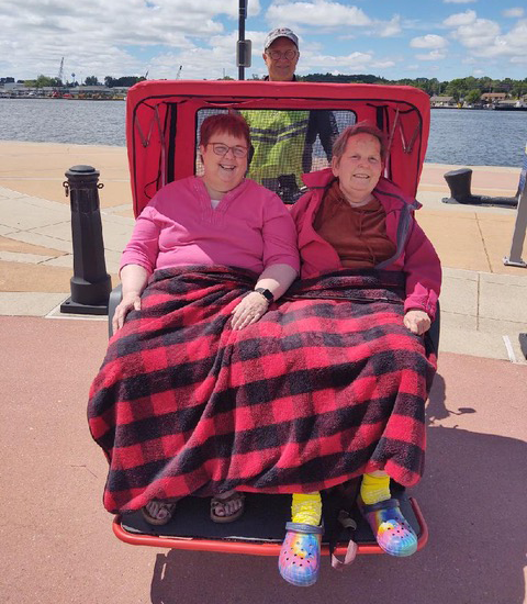 An elderly woman and a younger woman sitting in a Cyclign Without Age trishaw with a black and red checkered blanket covering both of them.