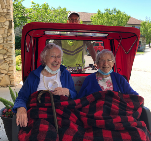 Two older people on a a Cycling Without Age trishaw. There is a black and red checkered blanket on both of them.