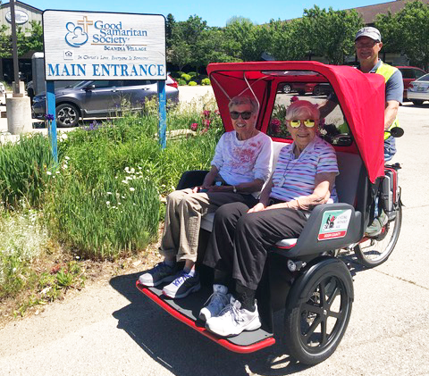 Two elderly people riing a trishaw with the pilot behind them. They are in front of a "Good Samaritan Society" sign.